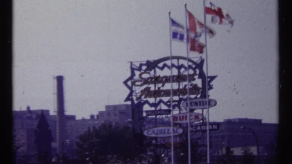 1967 Montreal frame showing a large Saraguay Automobiles sign, multiple flags, and distant buildings in soft 8mm color.