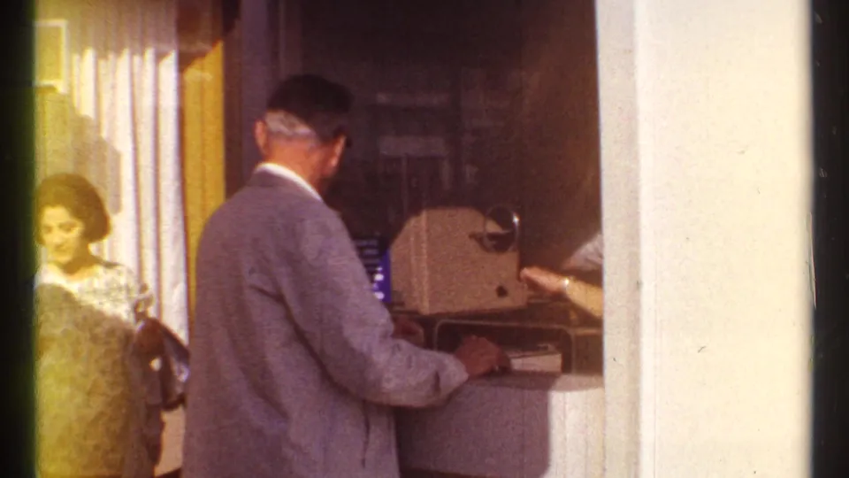 At the Montreal ticket booth, the suited man faces the counter while a woman in a patterned dress waits at the side.