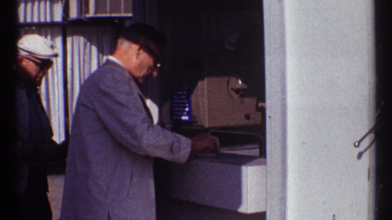 Men stand at a ticket counter beside a column, with one man using a machine.