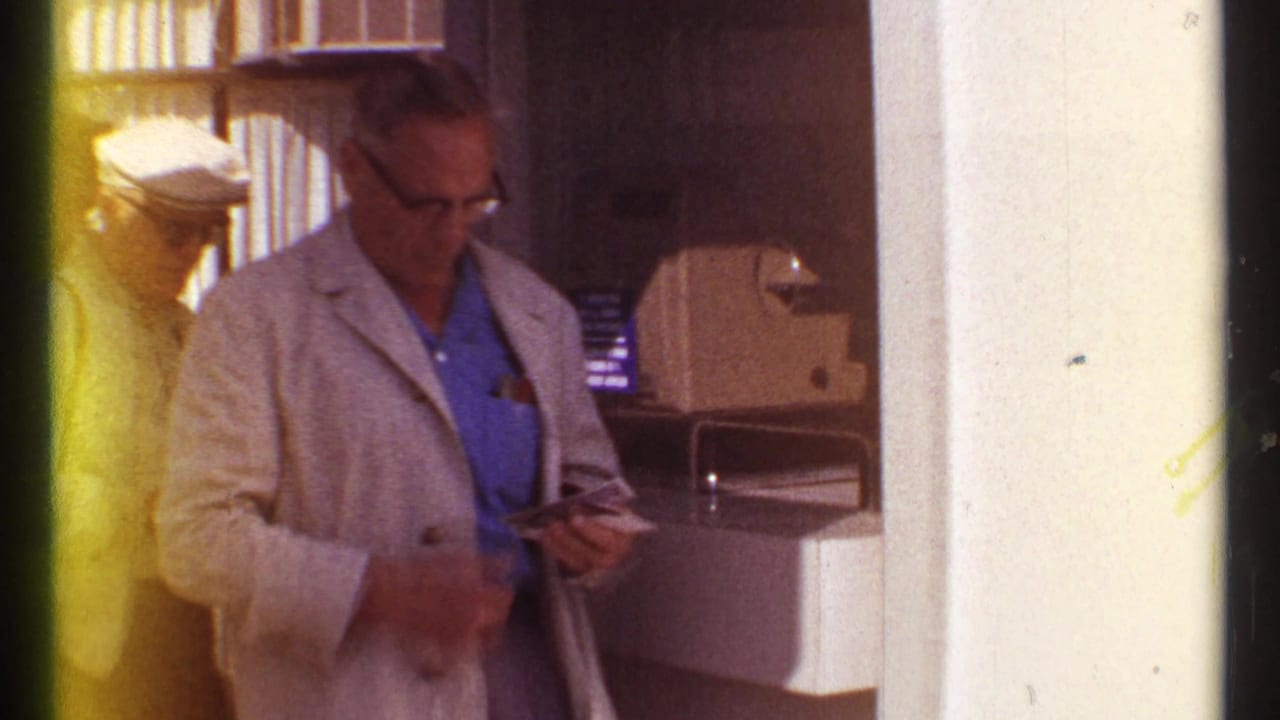 Man in a light jacket looks down at papers near a counter and column.