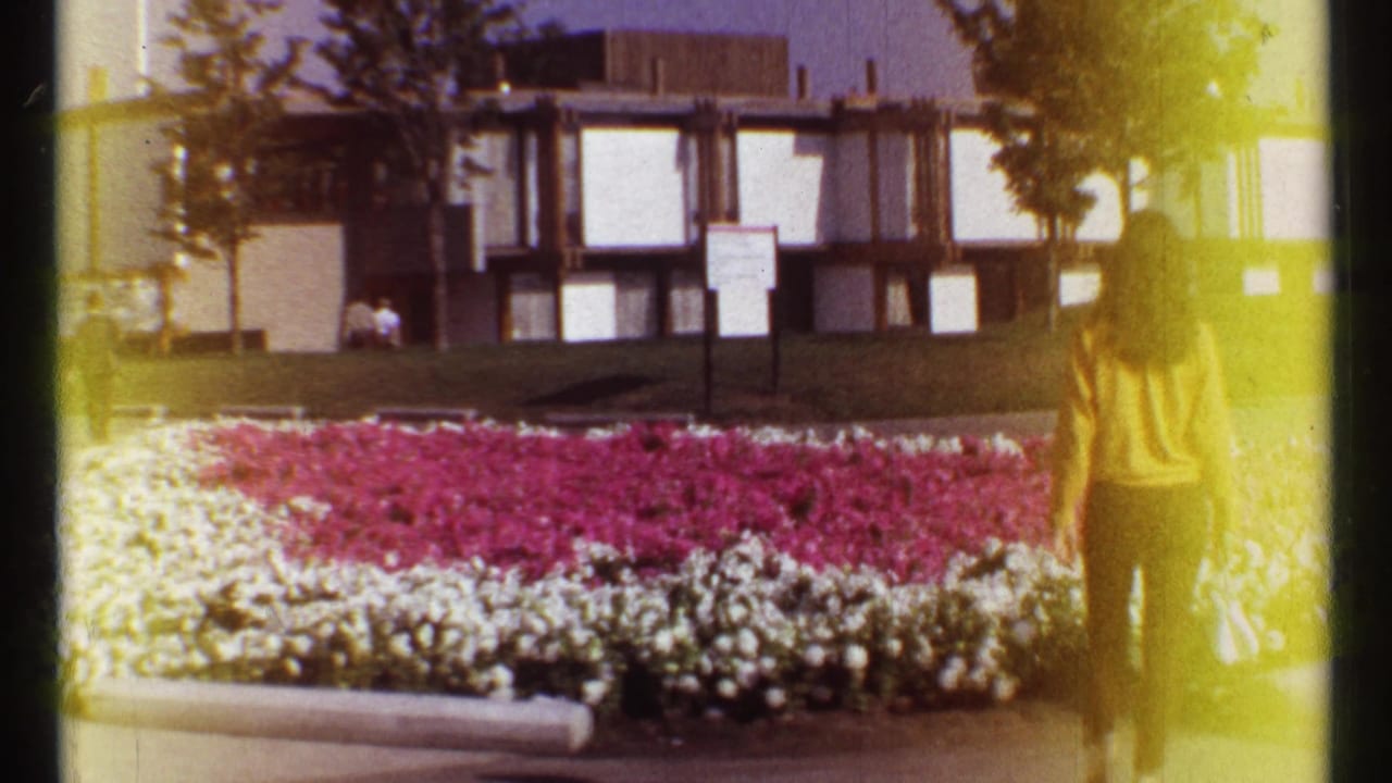 A flower bed sits in front of a modern building as a woman stands at the right edge.