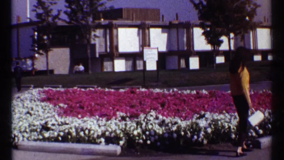 A woman in a yellow sweater stands near a vivid flower bed at the 1967 Montreal Expo, with a modern building and lawn behind her.