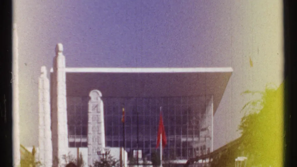 1967 Montreal 8mm frame showing a modern facade near Habitat 67, with vertical supports, glass, trees, and a red flag.