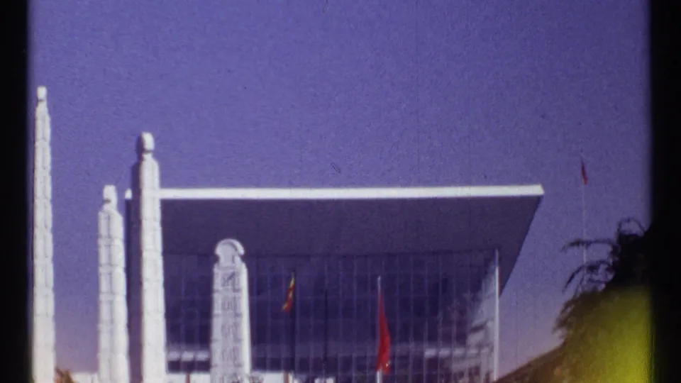 1967 Montreal 8mm frame showing a glass-fronted modernist building, white vertical pillars, flags, and a deep colored sky.