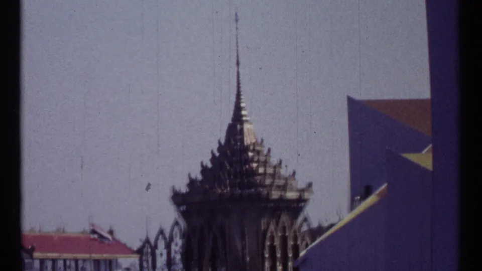 1967 Montreal 8mm frame holding on a dark traditional-style spire beside an angular wall and small flags under a washed pale sky.