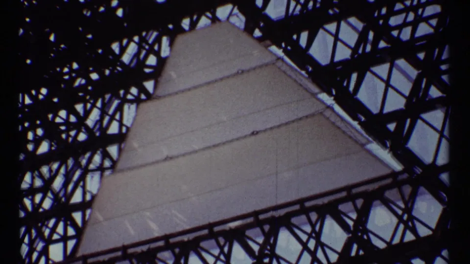 1967 Montreal home-movie frame showing pale monument panels seen through dark steel trusses from below.