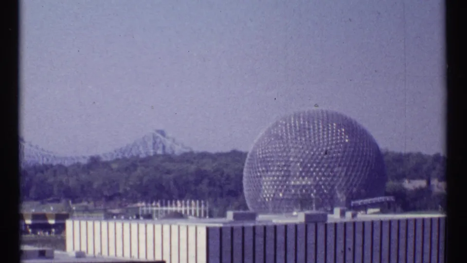 1967 Montreal 8mm view of a geodesic sphere beside low buildings, with trees and a distant bridge under a hazy sky.