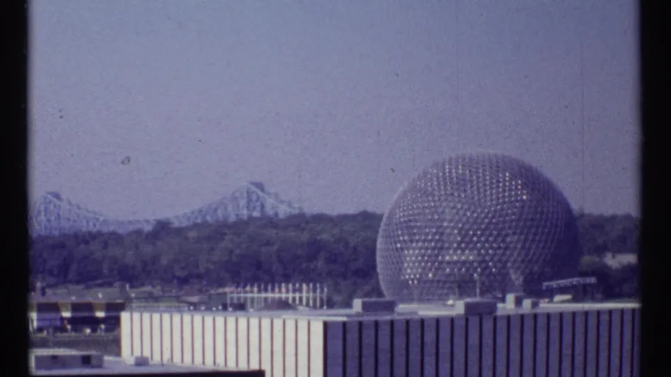 Wide 1967 Montreal home-movie frame showing the geodesic sphere, bridge, tree line, and pale buildings in soft 8mm color.