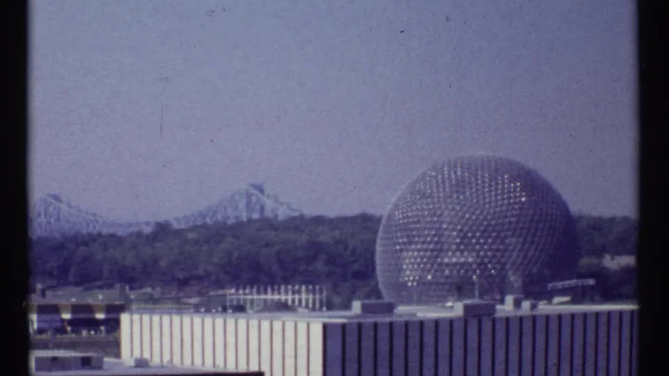 1967 Montreal Expo 67 view of a geodesic dome, distant roller coaster, trees, and low fairground buildings.