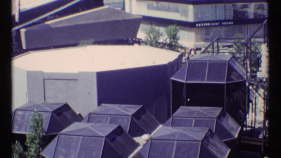 Visitor-shot 1967 Montreal Expo 67 view of dark polygonal roofs, a pale circular pavilion wall, and a stair or tower structure.