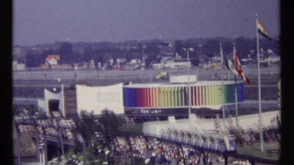 Wide 1967 Montreal Expo 67 view with a rainbow-striped pavilion facade, international flags, dense crowds, waterfront, and hazy hills beyond.