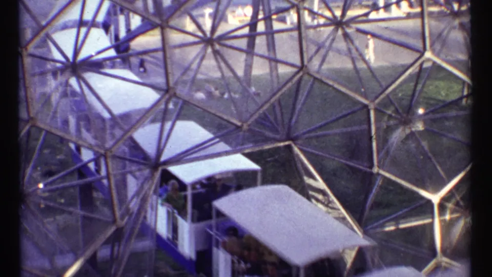 Visitors ride a small white-roofed train through the Montreal Expo dome interior, seen through angular structural panels.