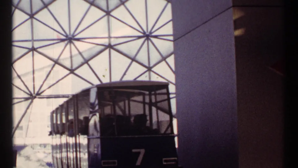 A side view of tram 7 shows seated passengers moving past the camera beneath the triangular station glazing in 1967 Montreal.