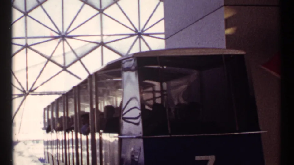 The tram fills the frame as it passes the camera, its dark passenger cabin set against the bright geodesic-pattern station wall.