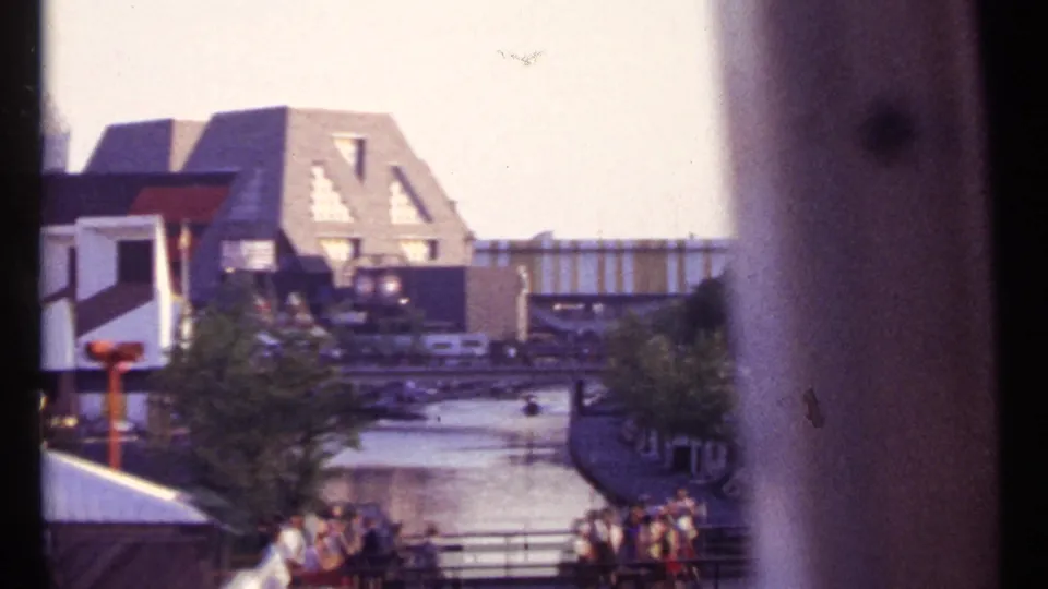 1967 8mm view of a Montreal waterway lined with pedestrians, trees, railings, and mid-century angular buildings.