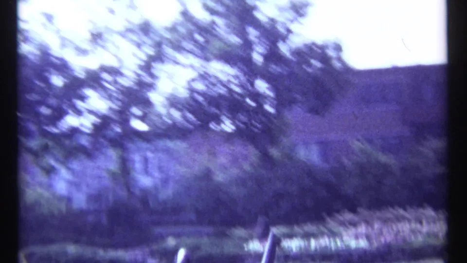 Motion-blurred 1967 Montreal 8mm frame showing trees, clipped hedges, a pale curved bench, and garden planting.
