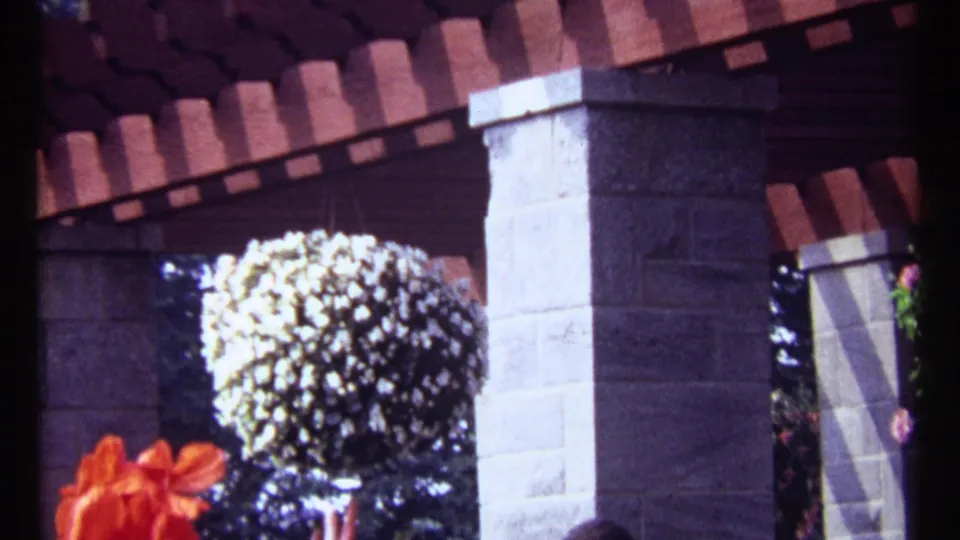 1967 Montreal 8mm frame of a stone-pillared pergola with hanging flowers, a large pale flowering basket, and brick roof details.