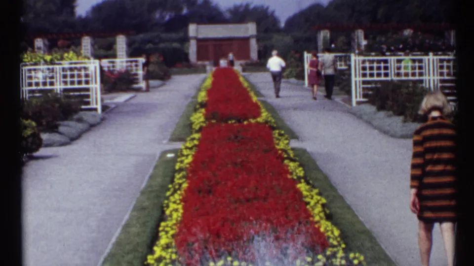 Wide 1967 Montreal garden view with visitors on formal paths, red flowerbeds bordered by yellow plantings, trellises, and brick garden buildings.