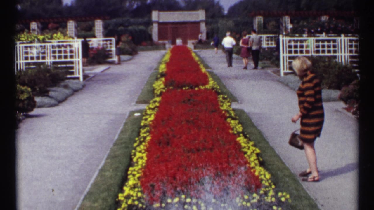 Woman in striped dress walks near a red flower bed as visitors continue along the path.