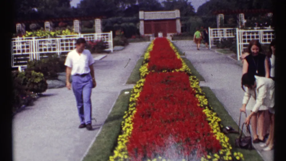 A man in a white shirt and blue trousers approaches the camera as other adults pause near the formal Montreal garden walkway.
