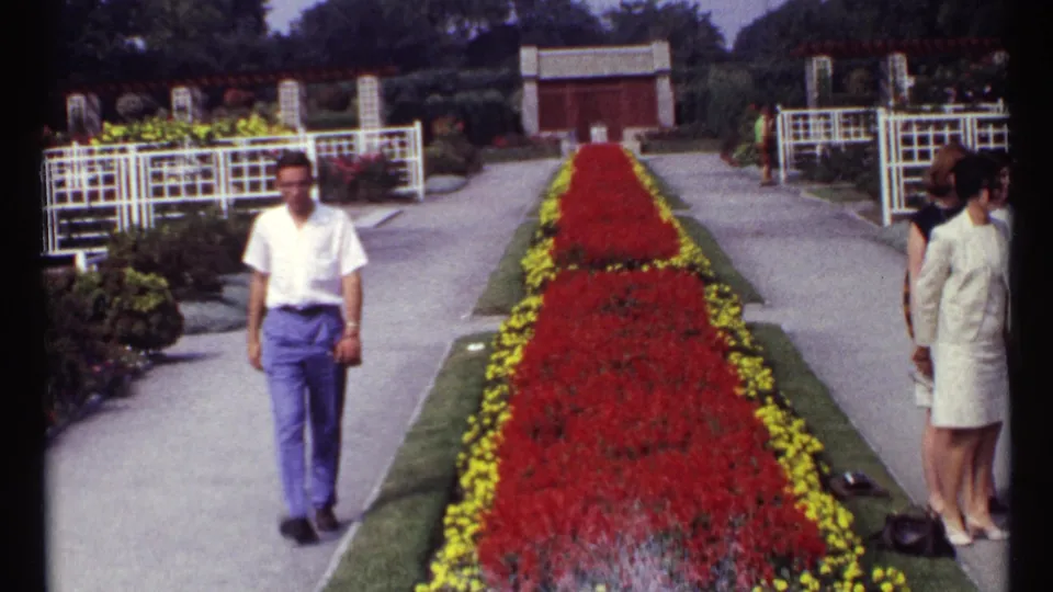 1967 Montreal home-movie frame showing a man walking through a formal flower garden as other adults stand near the path.