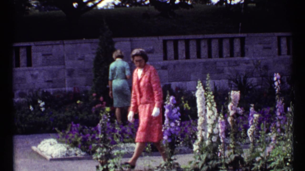 Women walking near flower beds in front of a low stone wall.