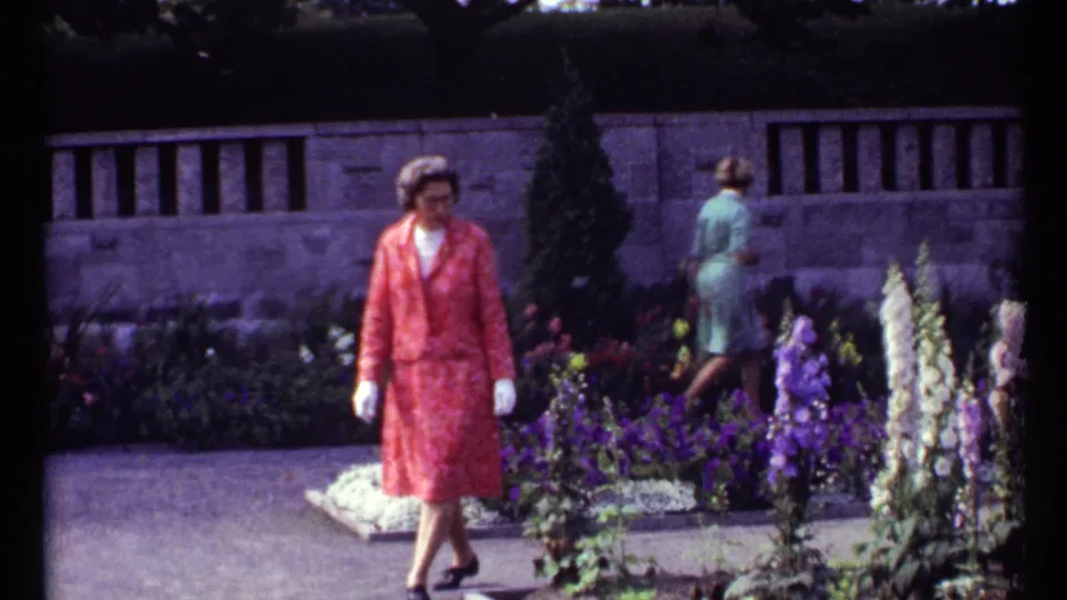 1967 Montreal home-movie frame of a woman in a coral skirt suit walking past planted beds while another visitor faces away.