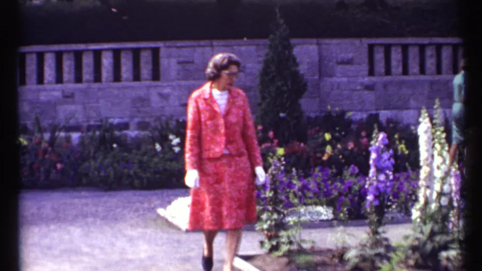 1967 Montreal garden frame showing a woman in a coral outfit near colorful flower beds and a low stone wall.