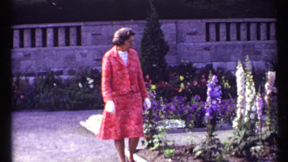 In 1967 Montreal footage, a woman in a red patterned dress and white gloves walks through a flower garden near a stone wall.