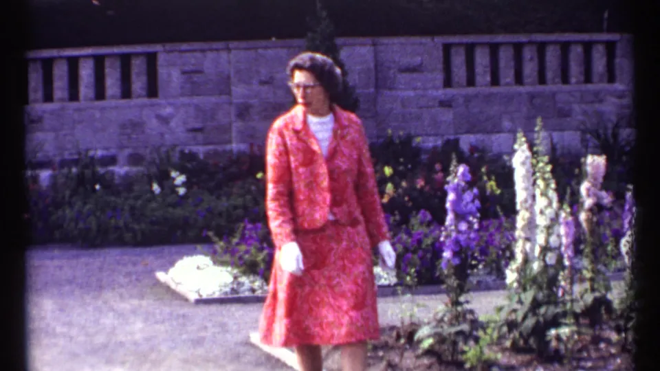 The woman in a red patterned dress is framed near foxglove-like purple and white flowers in the Montreal garden scene.
