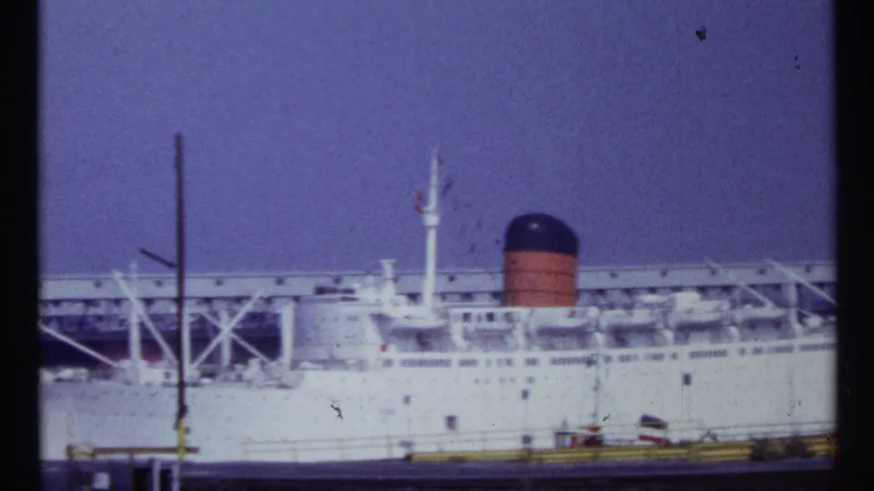 The docked passenger ship remains in view, showing its white hull, orange-and-black smokestack, pier framework, and hazy sky in Montreal footage.