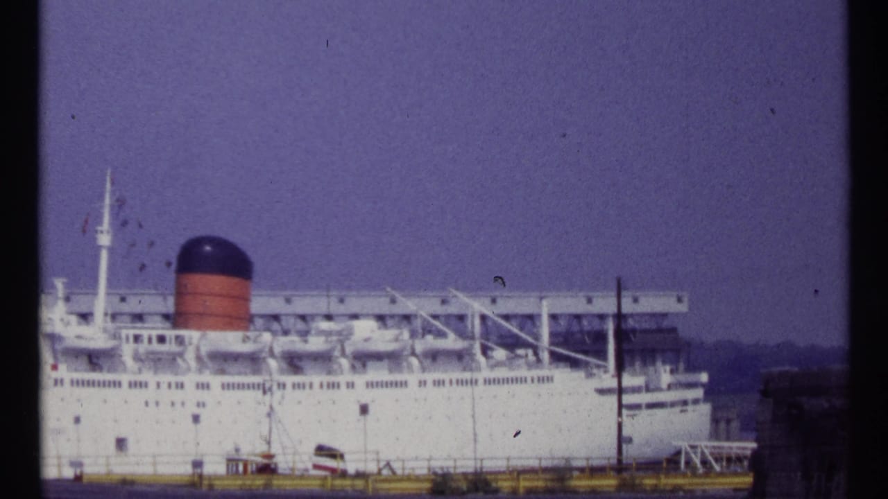 Docked passenger liner seen from shore with funnel, decks, lifeboats, and pier structures.