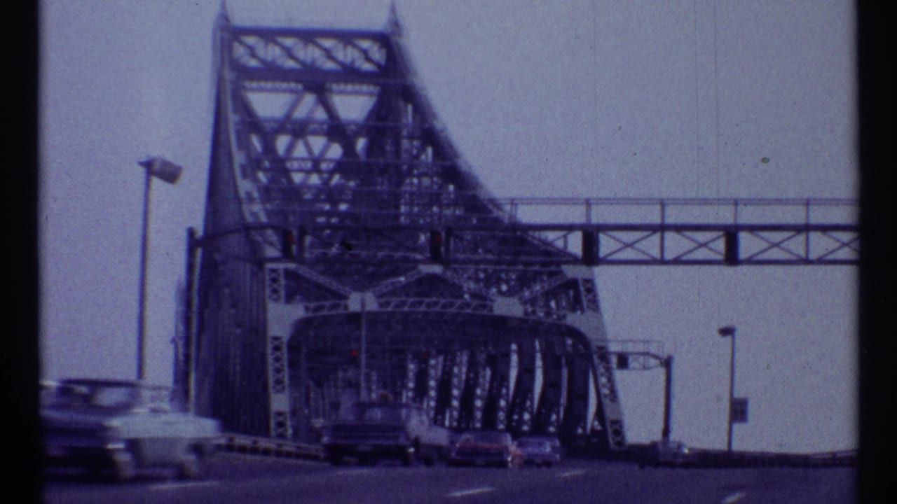 Moving roadway view toward a large steel truss bridge with cars ahead.