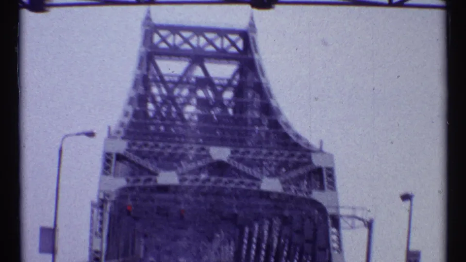 Forward-moving 1967 Montreal view beneath a steel bridge tower, with lamps and traffic signals visible near the roadway.