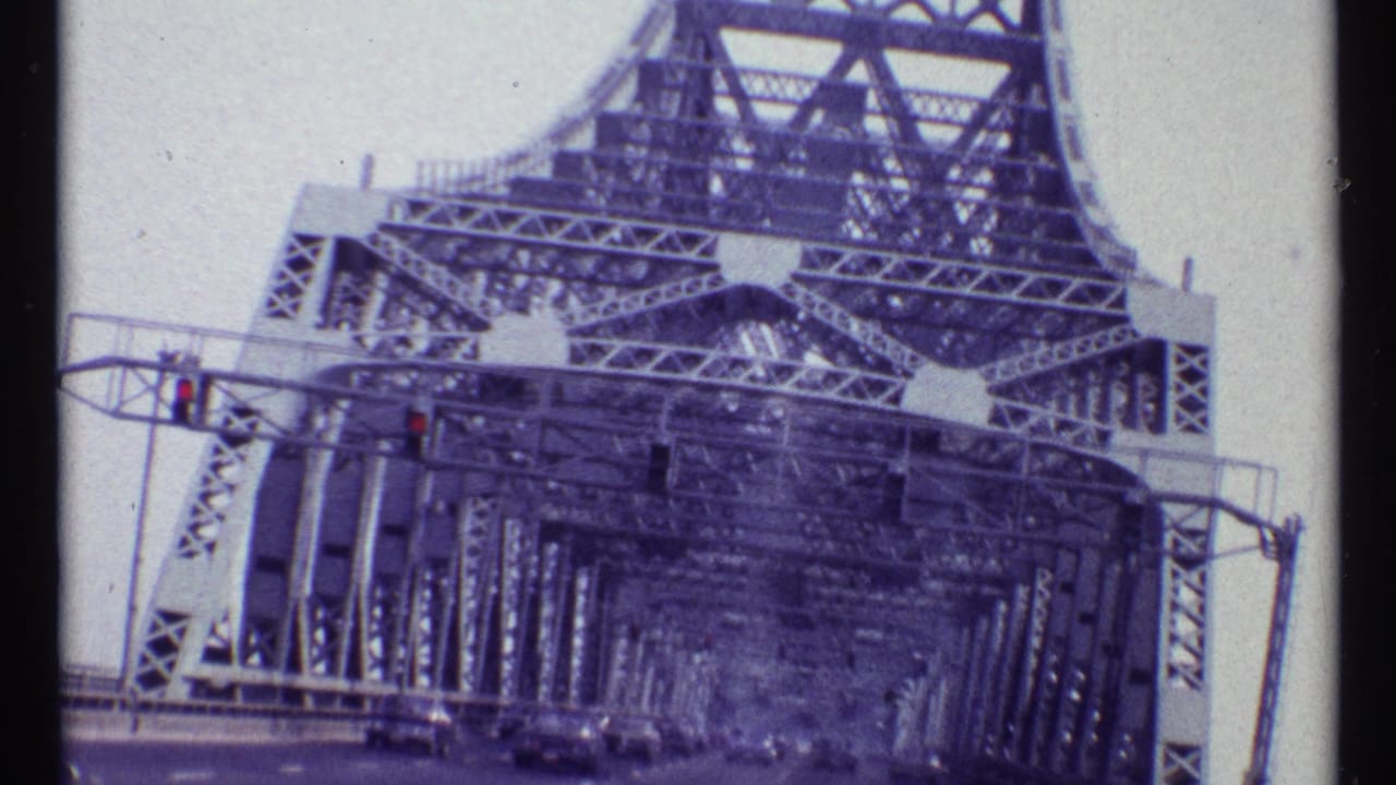 Low-angle roadway approach into a steel truss bridge under a pale cloudy sky.
