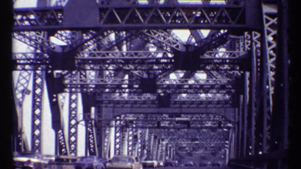 Inside the Alexandra Bridge structure in Montreal, the camera looks through repeating steel crossbeams toward cars ahead.