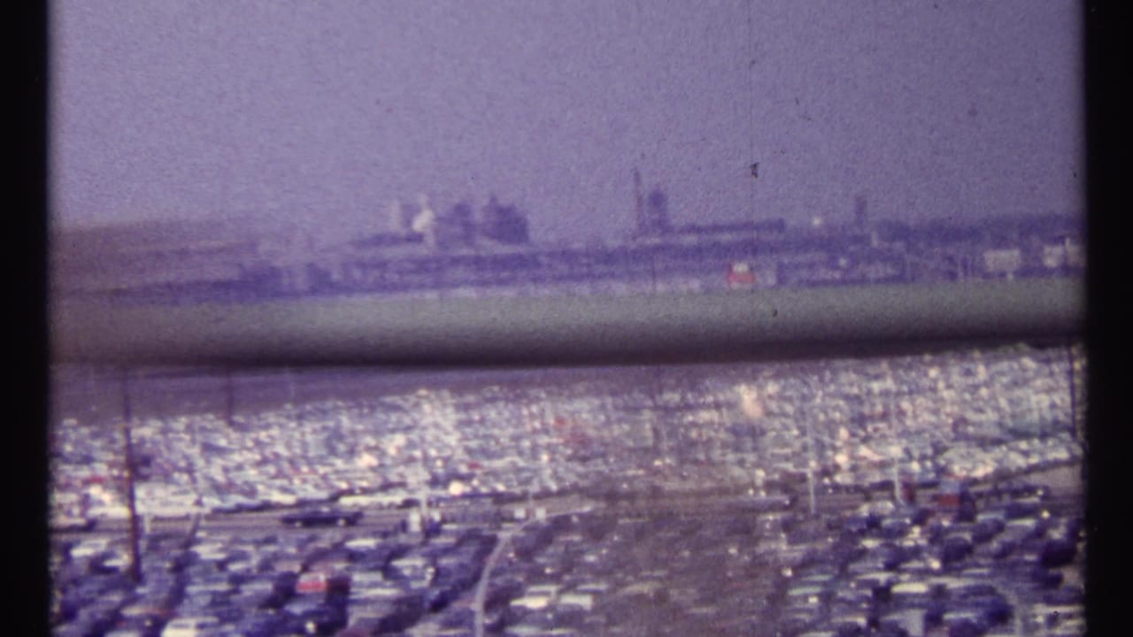 Crowded parking lot viewed past a horizontal railing with industrial structures beyond.