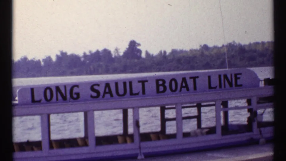 Close side view of the Long Sault Boat Line passenger boat, with readable lettering, windows, railings, water, and wooded shore.