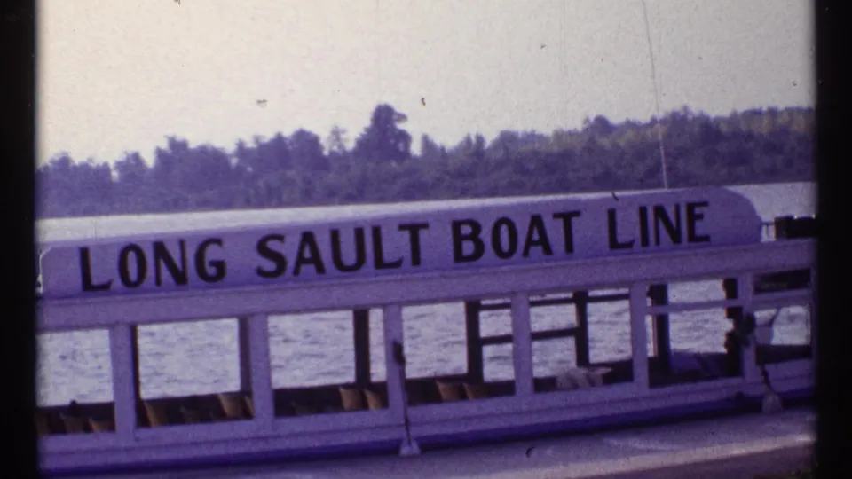 Later frame of the Long Sault Boat Line vessel on the river, with lettering, cabin windows, shoreline trees, and dark vertical framing.