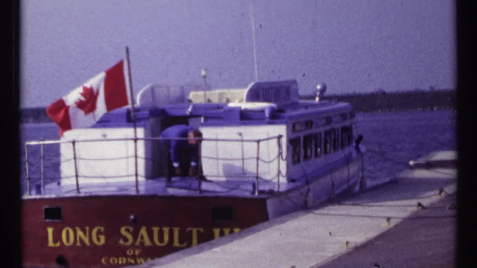 Home-movie frame from Montreal in 1967 showing the docked Long Sault boat, Canadian flag, and a man on deck.