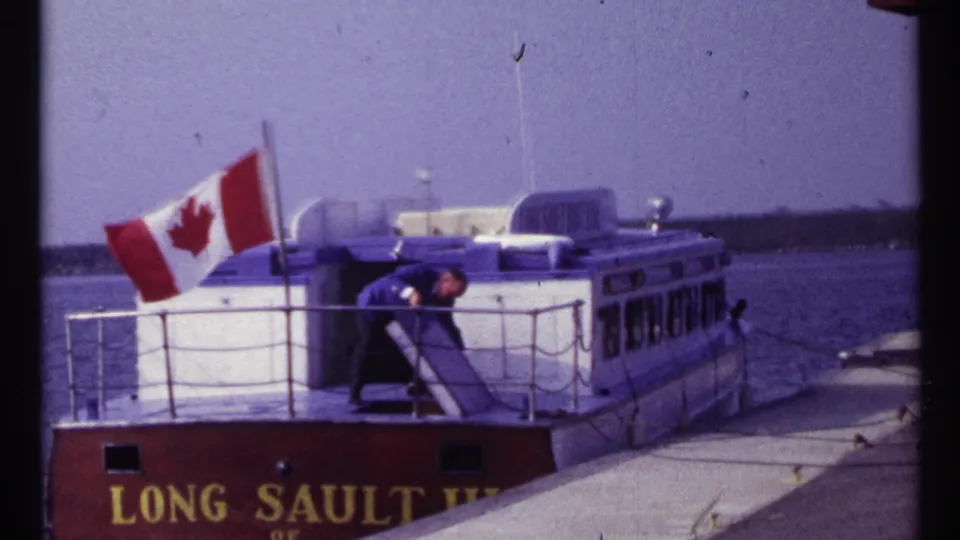 1967 Montreal 8mm still showing a man working on the stern of a Long Sault vessel with Canadian flag flying.