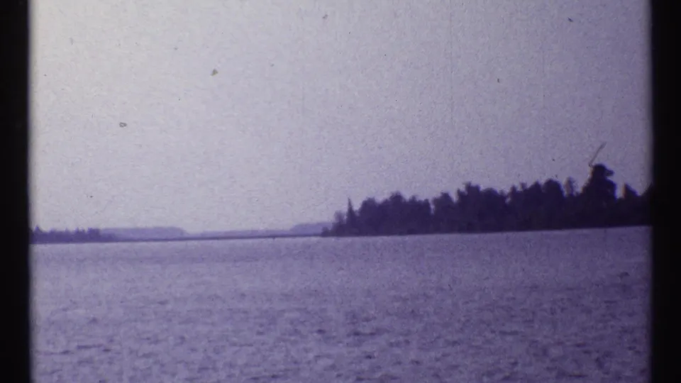 1967 8mm frame from the Montreal-area clip showing open water and a distant tree-lined shore under a pale hazy sky.