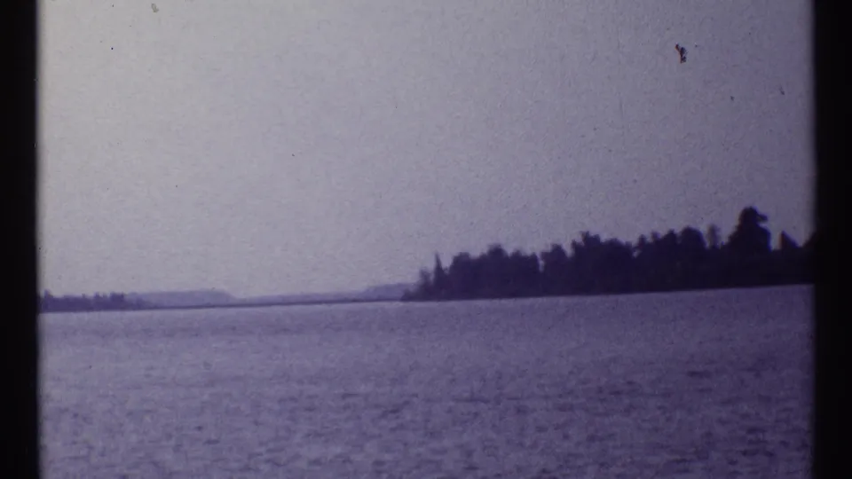 1960s 8mm home-movie frame showing lake water and a low wooded point, consistent with the Montreal-area boating clip metadata.