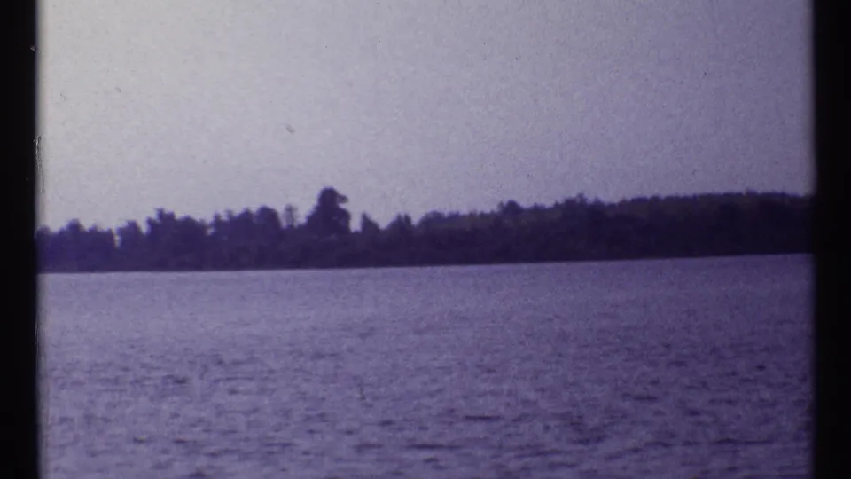 1967 Montreal 8mm frame showing open water, a dark tree-lined shoreline, and pale sky with purple film color cast.