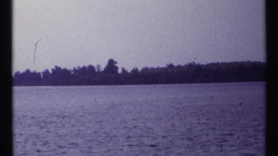 1967 Montreal home-movie frame of rippling water and a distant wooded shore, with soft focus and visible small-gauge film texture.