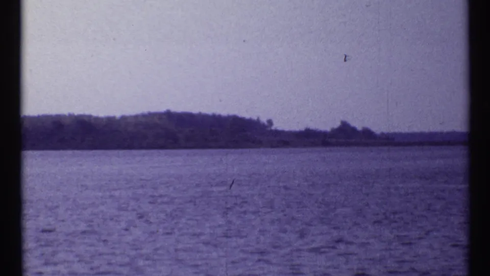 1967 Montreal 8mm frame showing a broad water surface and a low wooded shore across the background beneath a washed pale sky.