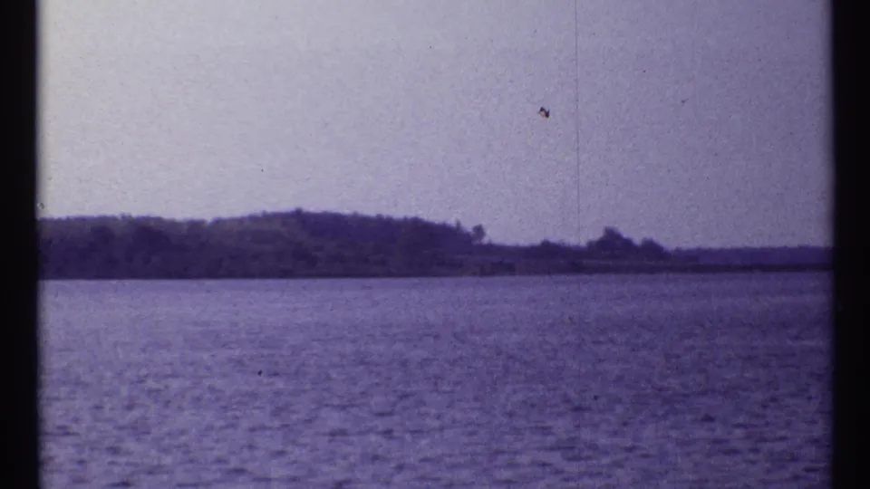 1967 Montreal home-movie frame of a calm body of water and distant tree-covered shore, tinted purple by the aged film scan.