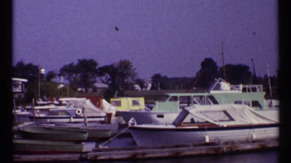 1967 Montreal home-movie frame of a quiet marina with moored boats, dock rails, trees, and calm water.