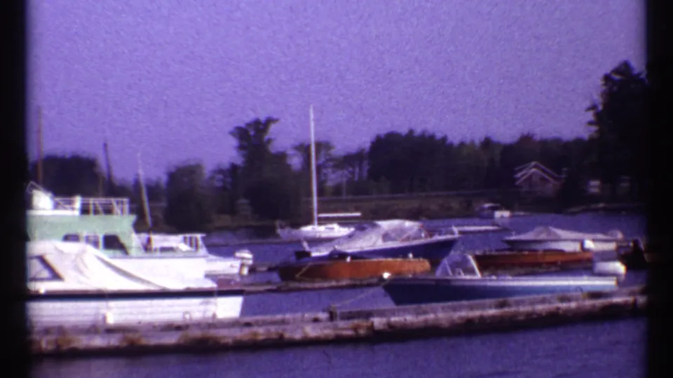 1967 Montreal 8mm frame showing covered small boats, a sailboat mast, dock edge, and wooded shoreline.