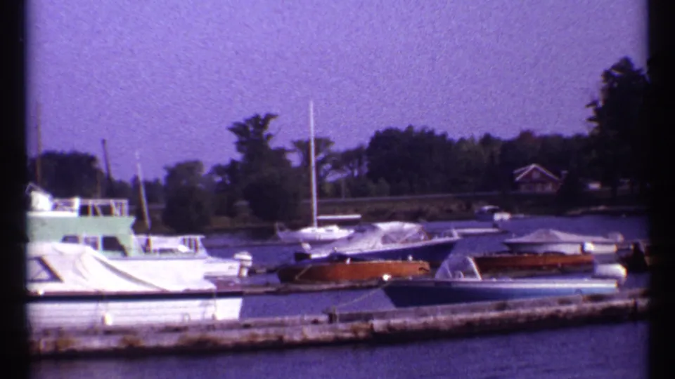 1967 Montreal home-movie still of moored covered boats along a dock, with trees and a small shoreline building behind.